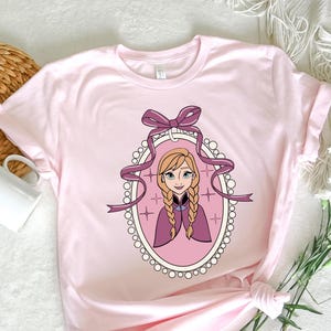 May include: A light pink t-shirt featuring a cartoon portrait of a character with blonde braids, wearing a purple cape, and framed by a purple ribbon and an oval border. The shirt is knotted at the bottom.