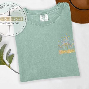 May include: A light green Comfort Colors t-shirt with an embroidered castle design and the word "Disneyland" in gold thread. The shirt also features a circular graphic that says "Personalized Gift" and "Embroidered Comfort Colors."