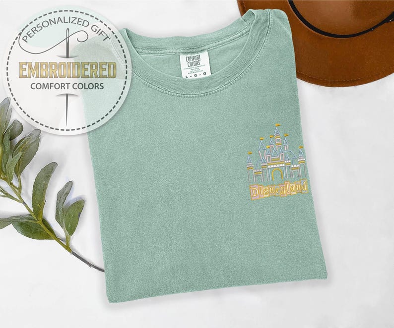 May include: A light green Comfort Colors t-shirt with an embroidered castle design and the word "Disneyland" in gold thread. The shirt also features a circular graphic that says "Personalized Gift" and "Embroidered Comfort Colors."