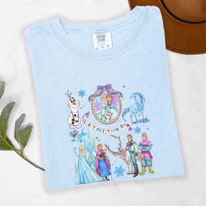 May include: Light blue t-shirt featuring a colorful graphic of characters from the movie Frozen, including Elsa, Anna, Olaf, and Kristoff. The words "LET IT GO" are also displayed. The shirt is made of a soft material and is a Comfort Colors brand.