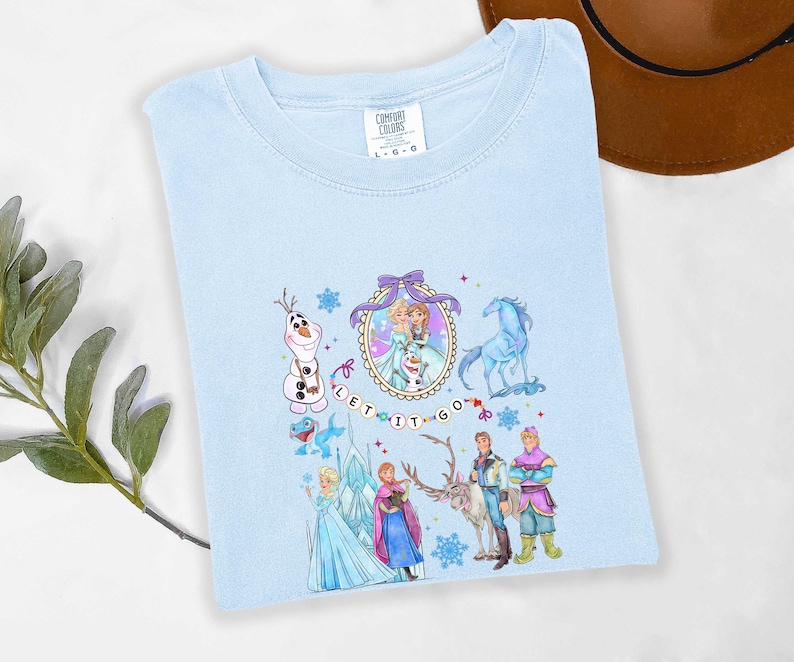 May include: Light blue t-shirt featuring a colorful graphic of characters from the movie Frozen, including Elsa, Anna, Olaf, and Kristoff. The words "LET IT GO" are also displayed. The shirt is made of a soft material and is a Comfort Colors brand.