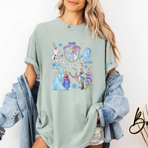 May include: A light green t-shirt featuring a colorful graphic of characters from the movie Frozen, including Elsa, Anna, Olaf, and Kristoff. The phrase "Let It Go" is also displayed. The shirt has rolled-up sleeves.