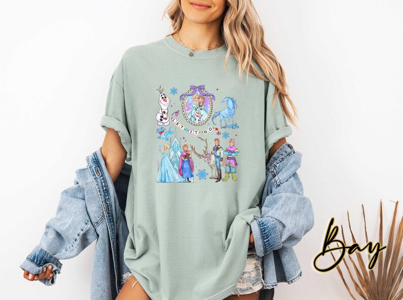 May include: A light green t-shirt featuring a colorful graphic of characters from the movie Frozen, including Elsa, Anna, Olaf, and Kristoff. The phrase "Let It Go" is also displayed. The shirt has rolled-up sleeves.