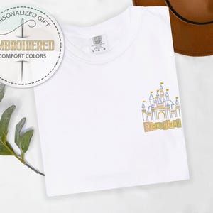 May include: White t-shirt with an embroidered Disneyland castle design in gold, blue, and yellow. The shirt also features the text "Disneyland" and a circular logo that says "Personalized Gift" and "Embroidered Comfort Colors".