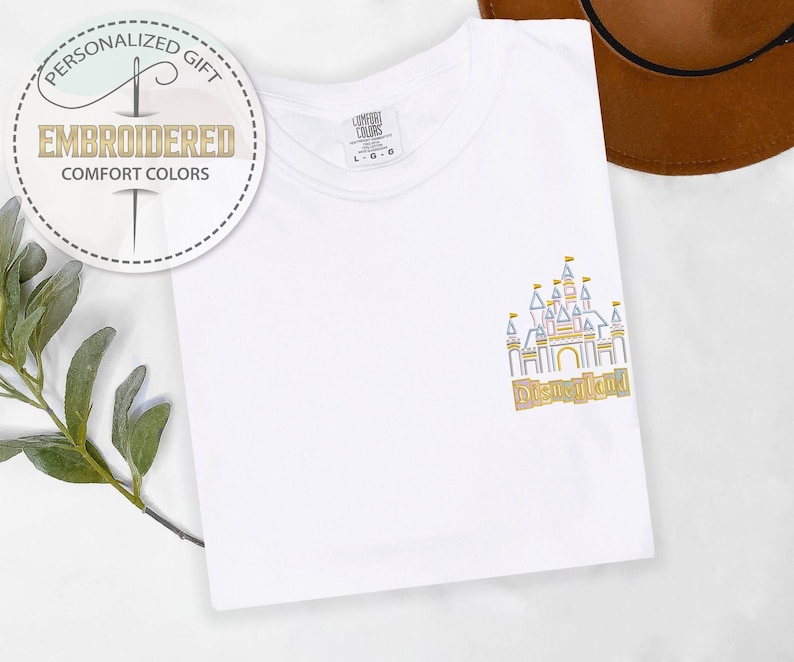May include: White t-shirt with an embroidered Disneyland castle design in gold, blue, and yellow. The shirt also features the text "Disneyland" and a circular logo that says "Personalized Gift" and "Embroidered Comfort Colors".