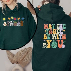 May include: Dark green hoodie featuring Star Wars characters and the text "May the Force Be With You." The front displays characters with Mickey Mouse balloon designs. The back has the phrase in colorful, retro lettering.