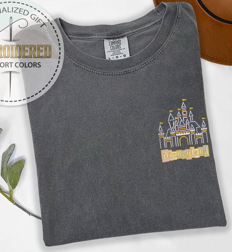 May include: A dark gray t-shirt with an embroidered Disneyland castle design in yellow, pink, and blue. The shirt has a crew neck and the Comfort Colors brand tag is visible. The text "Disneyland" is below the castle.