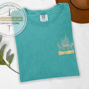 May include: Teal Comfort Colors t-shirt with an embroidered Disneyland castle design. The shirt features the text "Disneyland" in gold thread. A circular logo with the text "Personalized Gift" and "Embroidered Comfort Colors" is visible.