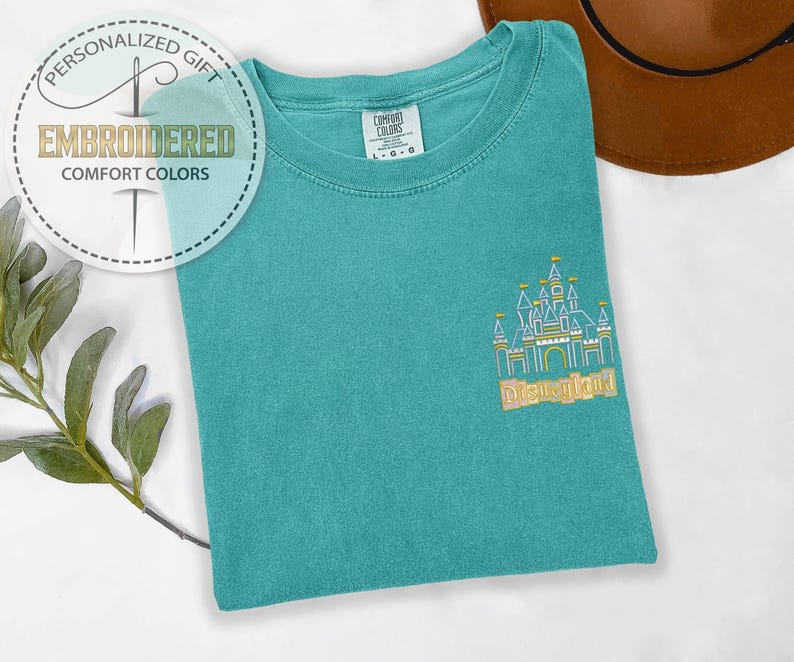 May include: Teal Comfort Colors t-shirt with an embroidered Disneyland castle design. The shirt features the text "Disneyland" in gold thread. A circular logo with the text "Personalized Gift" and "Embroidered Comfort Colors" is visible.
