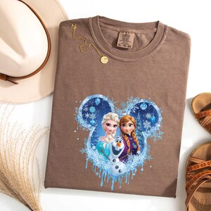 May include: A brown t-shirt featuring a Disney Frozen graphic with Elsa, Anna, and Olaf in a blue and white design. The shirt is paired with a beige hat, gold necklace, and brown sandals. The shirt size is XL.