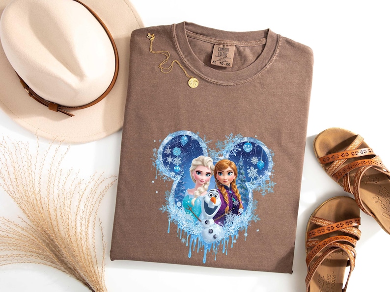 May include: A brown t-shirt featuring a Disney Frozen graphic with Elsa, Anna, and Olaf in a blue and white design. The shirt is paired with a beige hat, gold necklace, and brown sandals. The shirt size is XL.