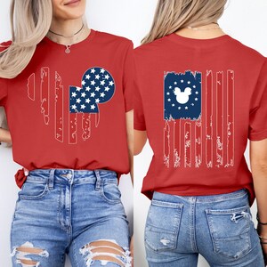 May include: Red t-shirt with a distressed American flag design incorporating a Mickey Mouse head silhouette. The front design is on the left, and the back design is centered. The flag's blue section has white stars. A casual, patriotic tee.