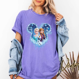 May include: A violet t-shirt featuring a graphic of Disney's Frozen characters within a Mickey Mouse silhouette. The design includes Elsa, Anna, and Olaf, with a blue and white icy theme. The shirt has short sleeves and a relaxed fit.