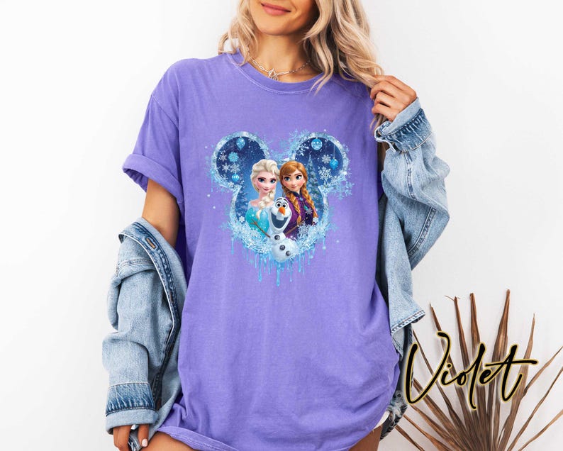 May include: A violet t-shirt featuring a graphic of Disney's Frozen characters within a Mickey Mouse silhouette. The design includes Elsa, Anna, and Olaf, with a blue and white icy theme. The shirt has short sleeves and a relaxed fit.