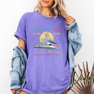 May include: Violet-colored t-shirt with a graphic of a monorail, castle, and text that reads "PLEASE STAND CLEAR OF THE DOORS" and "POR FAVOR, MANT&Eacute;NGASE ALEJADO DE LAS PUERTAS". The shirt has short sleeves.