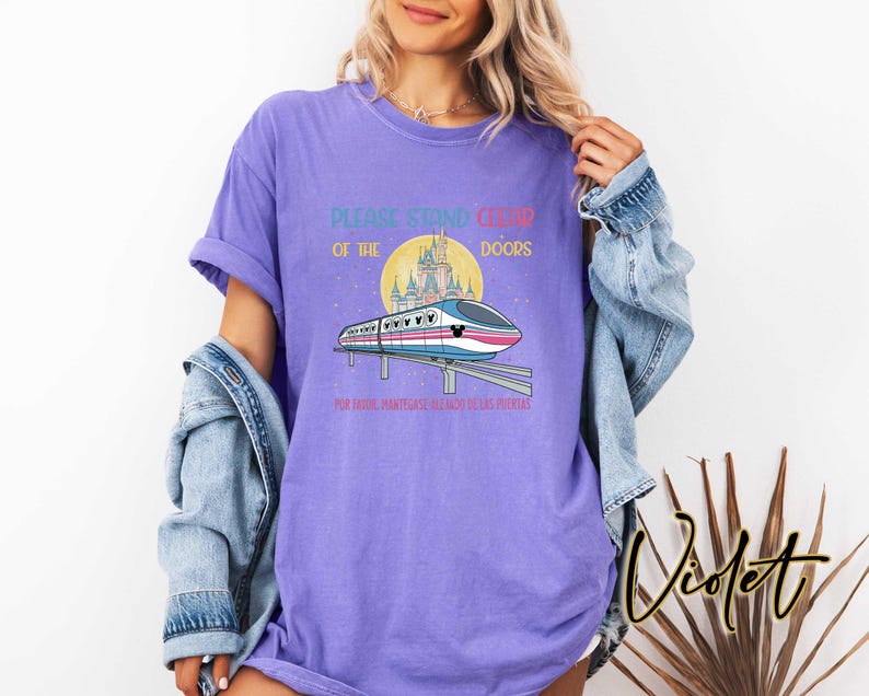 May include: Violet-colored t-shirt with a graphic of a monorail, castle, and text that reads "PLEASE STAND CLEAR OF THE DOORS" and "POR FAVOR, MANT&Eacute;NGASE ALEJADO DE LAS PUERTAS". The shirt has short sleeves.