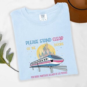 May include: Light blue t-shirt with the text "PLEASE STAND CLEAR OF THE DOORS" in colorful letters. The shirt features a cartoon train with a Mickey Mouse head design, a castle, and the Spanish phrase "POR FAVOR, MANT&Eacute;GASE ALEJADO DE LAS PUERTAS."