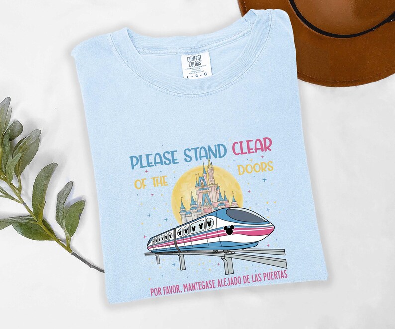 May include: Light blue t-shirt with the text "PLEASE STAND CLEAR OF THE DOORS" in colorful letters. The shirt features a cartoon train with a Mickey Mouse head design, a castle, and the Spanish phrase "POR FAVOR, MANT&Eacute;GASE ALEJADO DE LAS PUERTAS."