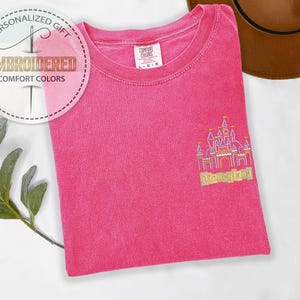 May include: A hot pink Comfort Colors t-shirt with an embroidered Disneyland castle design in gold and pink. The shirt has a crew neck and short sleeves. The image also includes a brown hat and a sprig of greenery.