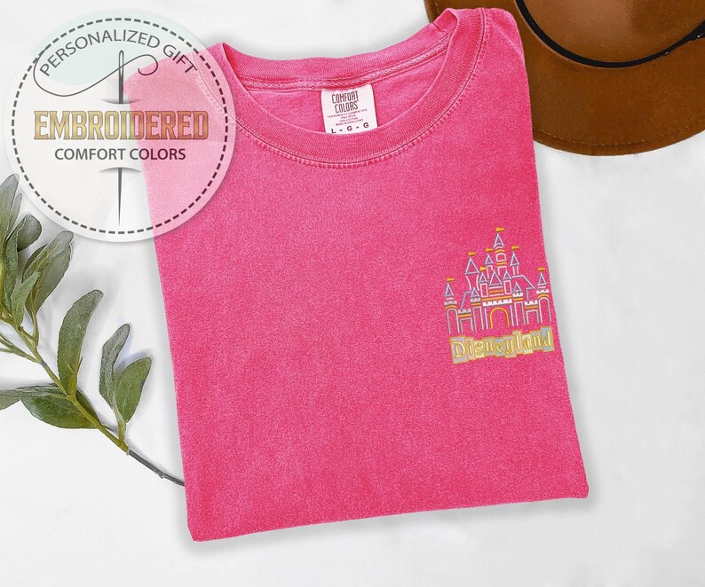 May include: A hot pink Comfort Colors t-shirt with an embroidered Disneyland castle design in gold and pink. The shirt has a crew neck and short sleeves. The image also includes a brown hat and a sprig of greenery.