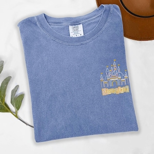 May include: A blue t-shirt with a gold embroidered image of a castle and the word "Disneyland". The shirt is a Comfort Colors brand and has a crew neck. A brown hat is visible in the background.