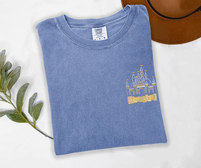 May include: A blue t-shirt with a gold embroidered image of a castle and the word "Disneyland". The shirt is a Comfort Colors brand and has a crew neck. A brown hat is visible in the background.
