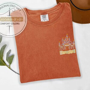 May include: Rust-colored Comfort Colors t-shirt with an embroidered Disneyland castle design. The shirt features the text "Embroidered Comfort Colors" and "Personalized Gift" in a circular logo. A brown hat is visible in the background.