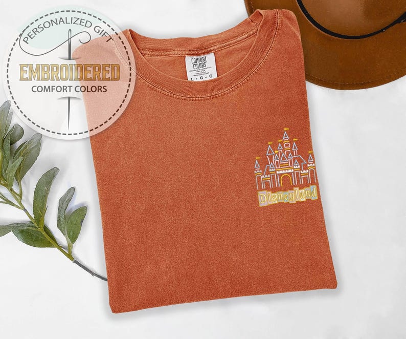 May include: Rust-colored Comfort Colors t-shirt with an embroidered Disneyland castle design. The shirt features the text "Embroidered Comfort Colors" and "Personalized Gift" in a circular logo. A brown hat is visible in the background.
