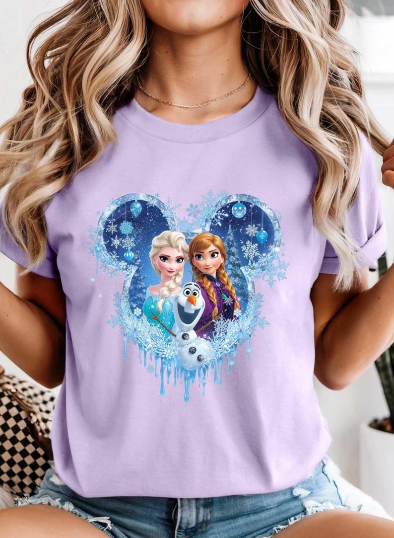May include: A light purple t-shirt featuring a graphic of Elsa, Anna, and Olaf from the movie Frozen. The design is set within a Mickey Mouse-shaped frame with snowflakes and icicles.