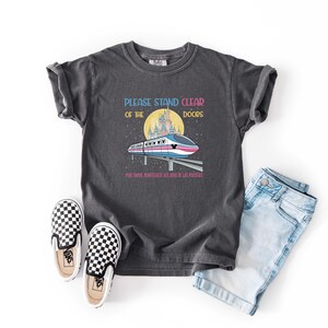 May include: Dark gray t-shirt with the text "PLEASE STAND CLEAR OF THE DOORS" and a graphic of a monorail in front of a castle. The shirt is paired with denim shorts and checkered slip-on shoes.