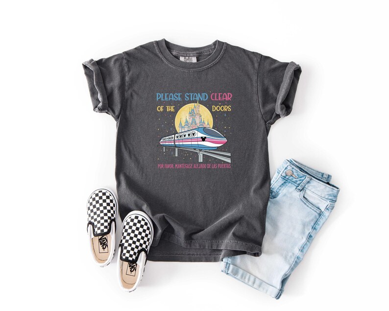 May include: Dark gray t-shirt with the text "PLEASE STAND CLEAR OF THE DOORS" and a graphic of a monorail in front of a castle. The shirt is paired with denim shorts and checkered slip-on shoes.