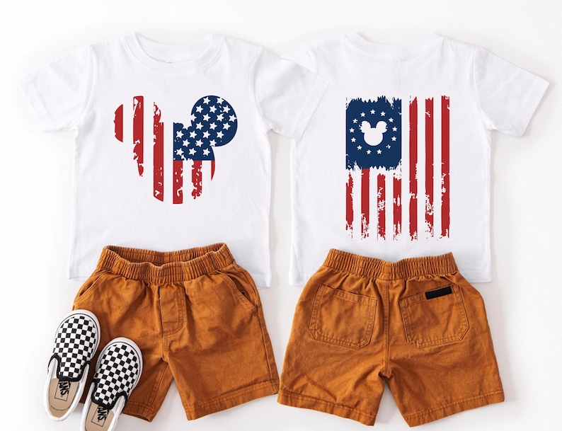 May include: Two white t-shirts with red, white, and blue American flag designs, one in the shape of a mouse head and the other a flag. Paired with orange shorts and black and white checkered slip-on shoes. A patriotic children's outfit.