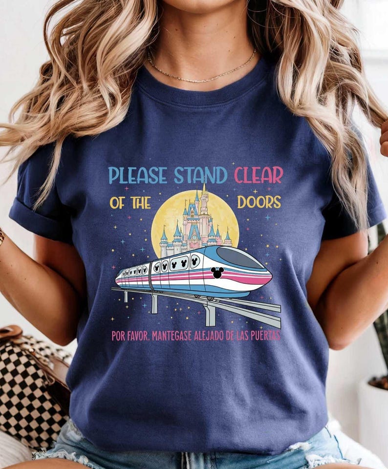 May include: Navy blue t-shirt featuring a monorail design with a castle and the text "PLEASE STAND CLEAR OF THE DOORS" in colorful letters. The design includes the text "POR FAVOR, MANTENGASE ALEJADO DE LAS PUERT" at the bottom.