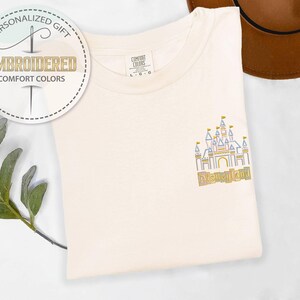May include: Cream-colored t-shirt with an embroidered Disneyland castle design in pastel colors. The shirt features the text "Disneyland" below the castle. The shirt also has a tag that says "Comfort Colors". A personalized gift.