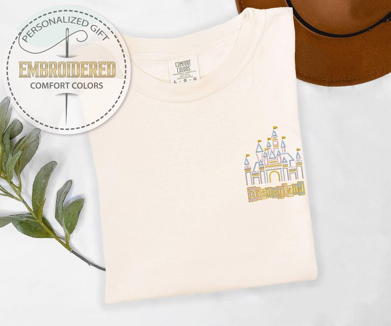 May include: Cream-colored t-shirt with an embroidered Disneyland castle design in pastel colors. The shirt features the text "Disneyland" below the castle. The shirt also has a tag that says "Comfort Colors". A personalized gift.