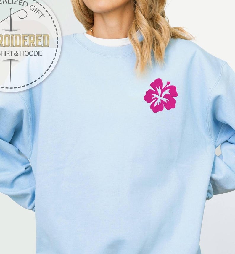 Puede incluir: Sudadera azul claro con una flor de hibisco fucsia bordada en el pecho. La sudadera tiene mangas largas y cuello redondo. La imagen tambi&eacute;n muestra un logotipo circular que dice "Regalo Personalizado" y "Camiseta y Sudadera con Bordado".