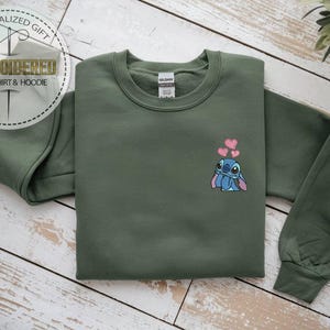 Puede incluir: Una sudadera verde oliva con un dise&ntilde;o de Stitch de dibujos animados y tres corazones rosas. La sudadera tiene mangas largas y cuello redondo. El texto "REGALO PERSONALIZADO" y "SUDADERA Y SUDADERA CON CAPUCHA BORDADA" est&aacute;n en un dise&ntilde;o circular.