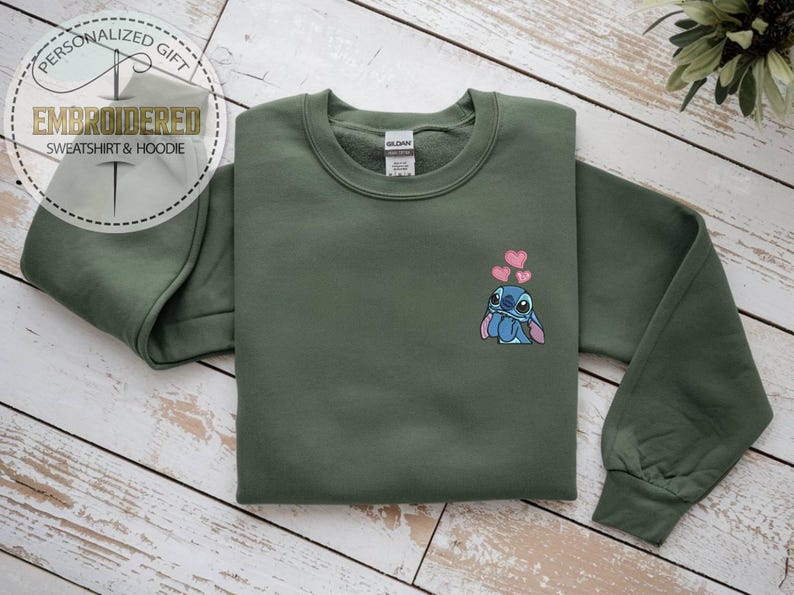 Puede incluir: Una sudadera verde oliva con un dise&ntilde;o de Stitch de dibujos animados y tres corazones rosas. La sudadera tiene mangas largas y cuello redondo. El texto "REGALO PERSONALIZADO" y "SUDADERA Y SUDADERA CON CAPUCHA BORDADA" est&aacute;n en un dise&ntilde;o circular.