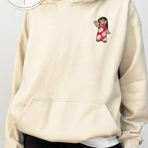 Puede incluir: Sudadera con capucha bordada de color crema con bolsillo delantero y capucha con cord&oacute;n. El dise&ntilde;o presenta un personaje de dibujos animados con un vestido rojo con lunares blancos. El texto "PERSONALIZED GIFT EMBROIDERED SWEATSHIRT & HOODIE" est&aacute; impreso arriba a la izquierda.