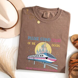 May include: Brown t-shirt with a graphic of a monorail, castle, and the text "PLEASE STAND CLEAR OF THE DOORS" in blue, pink, and yellow. The shirt also has text in Spanish. A gold necklace, sandals, and a hat are also in the image.