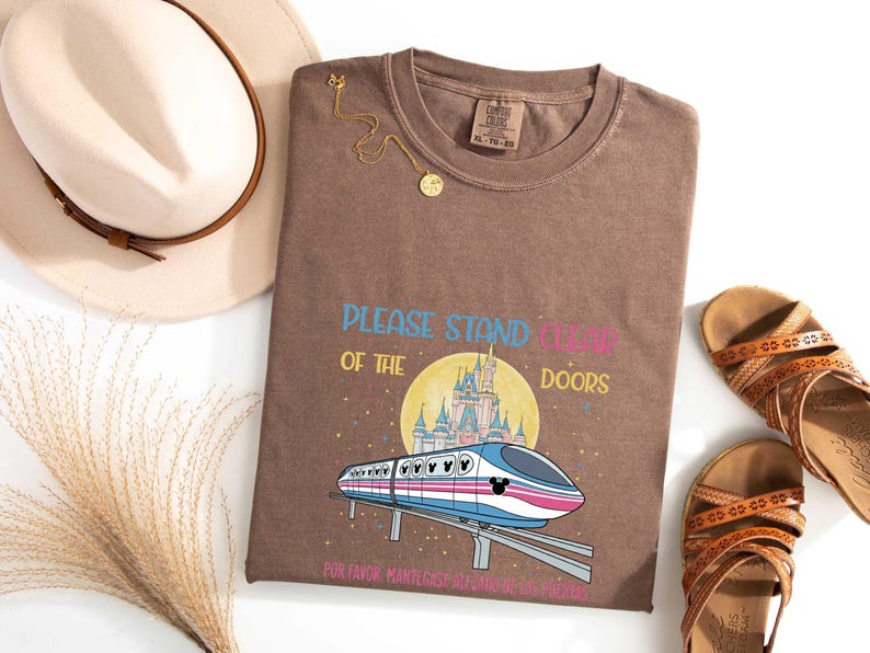 May include: Brown t-shirt with a graphic of a monorail, castle, and the text "PLEASE STAND CLEAR OF THE DOORS" in blue, pink, and yellow. The shirt also has text in Spanish. A gold necklace, sandals, and a hat are also in the image.