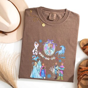May include: Brown t-shirt featuring a colorful graphic of characters from a popular animated movie. The design includes Elsa, Anna, Olaf, and other characters, with the words "Let it Go". The shirt is styled with a hat, sandals, and a necklace.
