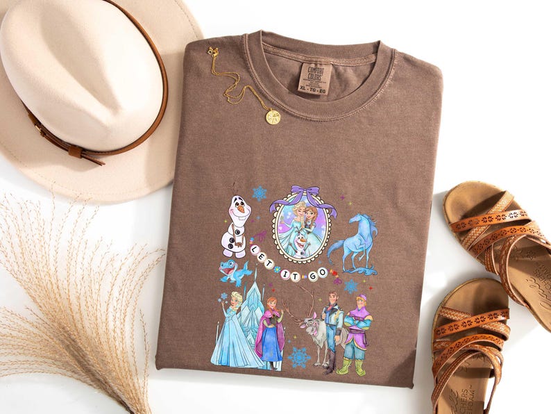May include: Brown t-shirt featuring a colorful graphic of characters from a popular animated movie. The design includes Elsa, Anna, Olaf, and other characters, with the words "Let it Go". The shirt is styled with a hat, sandals, and a necklace.