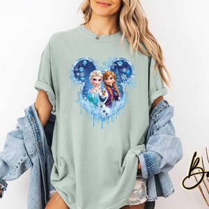 May include: A light green t-shirt featuring a graphic of Elsa, Anna, and Olaf from Disney's Frozen, set within a blue Mickey Mouse-shaped design with snowflakes. The shirt has rolled-up sleeves and a relaxed fit.