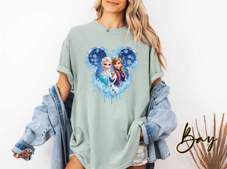 May include: A light green t-shirt featuring a graphic of Elsa, Anna, and Olaf from Disney's Frozen, set within a blue Mickey Mouse-shaped design with snowflakes. The shirt has rolled-up sleeves and a relaxed fit.