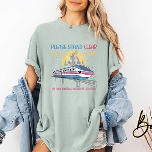 May include: A light green t-shirt with a graphic of a monorail in front of a castle and a yellow sun. The text on the shirt reads "PLEASE STAND CLEAR OF THE DOORS" and "POR FAVOR, MANT&Eacute;NGASE ALEJADO DE LAS PUERTAS".