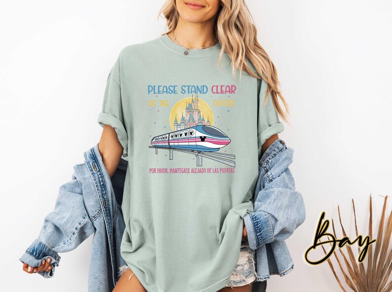 May include: A light green t-shirt with a graphic of a monorail in front of a castle and a yellow sun. The text on the shirt reads "PLEASE STAND CLEAR OF THE DOORS" and "POR FAVOR, MANT&Eacute;NGASE ALEJADO DE LAS PUERTAS".