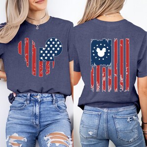 May include: Navy blue t-shirt with a distressed American flag design incorporating a Mickey Mouse silhouette. The front features a Mickey head shape with red stripes and a blue circle filled with white stars. The back has a flag design with a Mickey head.