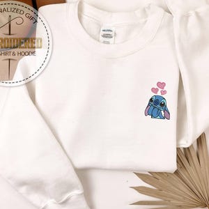 Puede incluir: Sudadera blanca con un dise&ntilde;o bordado de Stitch y corazones rosas. La sudadera tiene mangas largas y cuello redondo. La imagen tambi&eacute;n incluye un logotipo circular que dice "Regalo personalizado" y "Sudadera y sudadera con capucha bordadas".