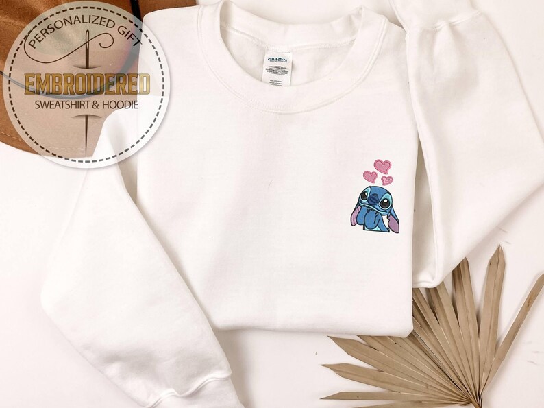 Puede incluir: Sudadera blanca con un dise&ntilde;o bordado de Stitch y corazones rosas. La sudadera tiene mangas largas y cuello redondo. La imagen tambi&eacute;n incluye un logotipo circular que dice "Regalo personalizado" y "Sudadera y sudadera con capucha bordadas".